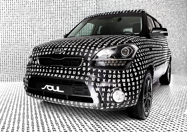 A black car covered in white letters arranged like a word search sits in a white room covered in black word search letters