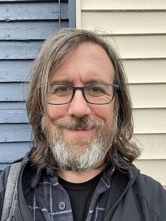 A head and shoulders portrait of a man with a beard and long hair, standing in front of a building with horizontal slats painted gray on the left and cream on the right.