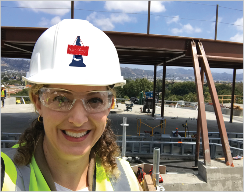 Woman on construction site with steel girders; she's wearing hardhat and safety goggles and fluorescent vest