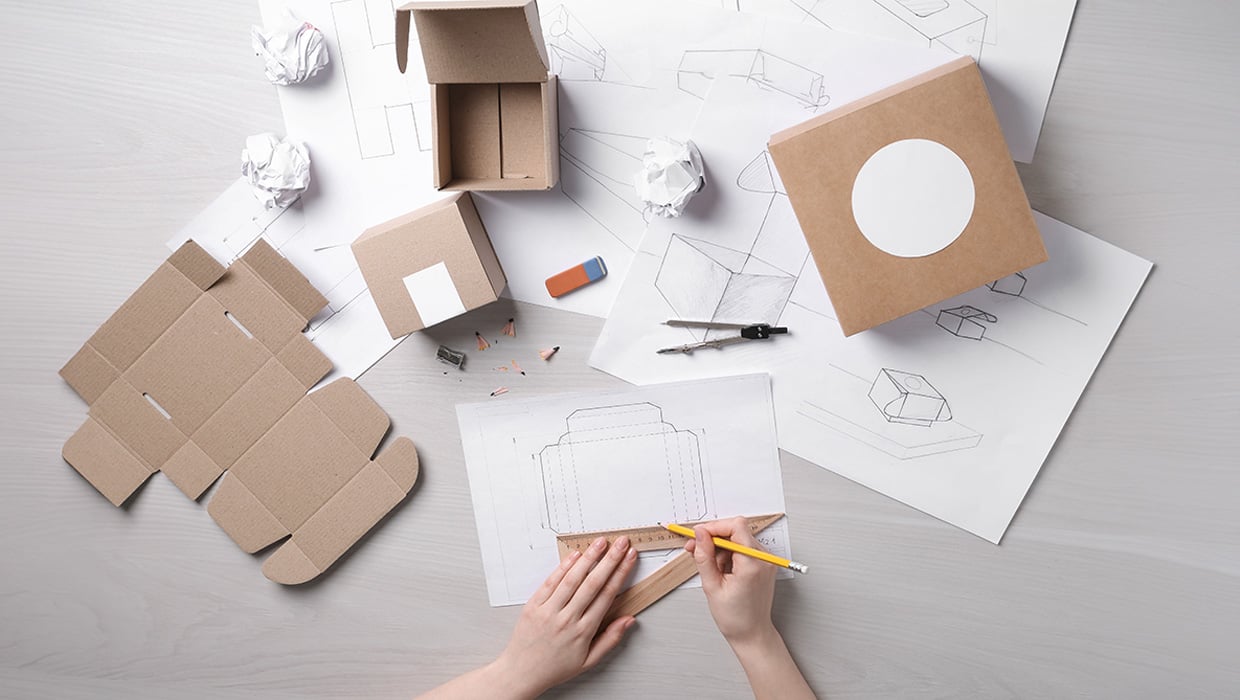 Overhead view of a packaging design desk with cardboard boxes, sketches, and a hand drawing a box layout.